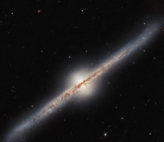 NASA’s Hubble Snaps Stunning Edge-On Spiral Galaxy Image A spiral galaxy seen directly from the side, such that its disk looks like a narrow diagonal band across the image. A band of dark dust covers the disk in the center most of the way out to the ends, and the disk glows around that. In the center of the galaxy, a whitish circle of light bulges out above and below the disk. Each end of the disk curves slightly. The background is black and mostly empty.