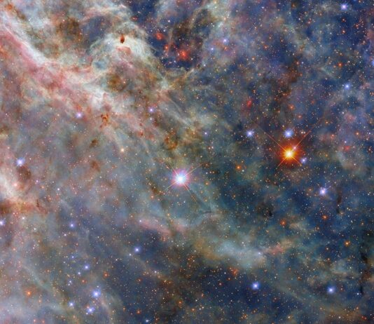 NASA’s Hubble Telescope Photographs Tarantula Nebula A colorful nebula. The image’s top-left is filled with layers of fluffy pink and greenish clouds. Long strands of green clouds stretch out from here; a faint layer of translucent blue dust combines with them to create a three-dimensional scene. A sparse network of dark dust clouds in the foreground adds reddish-black patches atop the nebula. Blue-white and orange stars, from our galaxy and beyond, are spread throughout the clouds.