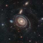 Hubble Explores Unique Nine-Ring Galaxy – NASA Science A large galaxy is at center, and a significantly smaller galaxy is to its left. The large galaxy, nicknamed the Bullseye, is mostly face-on, but the top appears slightly tilted away. It has several rings. Its circular core is bright white at the very center, but light yellow overall. Going outward, there are gaps between the rings. The core is surrounded by two slightly lighter yellow rings, which also appear to be overlapping. The next ring is slightly more transparent and yellow. The two or three rings that are farther out are bluer, sometimes with blue clumps. The widest ring is also blue, but also the most transparent. At 9 o’clock is a small dwarf galaxy. It is about the same size as the yellow core of the Bullseye. The dwarf galaxy is blue, with many dots. It looks like the edge of the Bullseye might touch the dwarf galaxy. Both galaxies are set on the black background of space, which is dotted with a range of galaxies in different shapes, colors, and sizes, along with one foreground star at left.