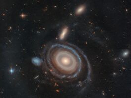 Hubble Explores Unique Nine-Ring Galaxy – NASA Science A large galaxy is at center, and a significantly smaller galaxy is to its left. The large galaxy, nicknamed the Bullseye, is mostly face-on, but the top appears slightly tilted away. It has several rings. Its circular core is bright white at the very center, but light yellow overall. Going outward, there are gaps between the rings. The core is surrounded by two slightly lighter yellow rings, which also appear to be overlapping. The next ring is slightly more transparent and yellow. The two or three rings that are farther out are bluer, sometimes with blue clumps. The widest ring is also blue, but also the most transparent. At 9 o’clock is a small dwarf galaxy. It is about the same size as the yellow core of the Bullseye. The dwarf galaxy is blue, with many dots. It looks like the edge of the Bullseye might touch the dwarf galaxy. Both galaxies are set on the black background of space, which is dotted with a range of galaxies in different shapes, colors, and sizes, along with one foreground star at left.