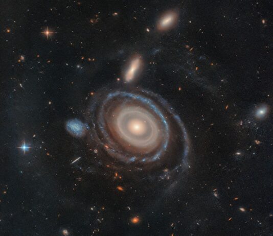 Hubble Explores Unique Nine-Ring Galaxy – NASA Science A large galaxy is at center, and a significantly smaller galaxy is to its left. The large galaxy, nicknamed the Bullseye, is mostly face-on, but the top appears slightly tilted away. It has several rings. Its circular core is bright white at the very center, but light yellow overall. Going outward, there are gaps between the rings. The core is surrounded by two slightly lighter yellow rings, which also appear to be overlapping. The next ring is slightly more transparent and yellow. The two or three rings that are farther out are bluer, sometimes with blue clumps. The widest ring is also blue, but also the most transparent. At 9 o’clock is a small dwarf galaxy. It is about the same size as the yellow core of the Bullseye. The dwarf galaxy is blue, with many dots. It looks like the edge of the Bullseye might touch the dwarf galaxy. Both galaxies are set on the black background of space, which is dotted with a range of galaxies in different shapes, colors, and sizes, along with one foreground star at left.