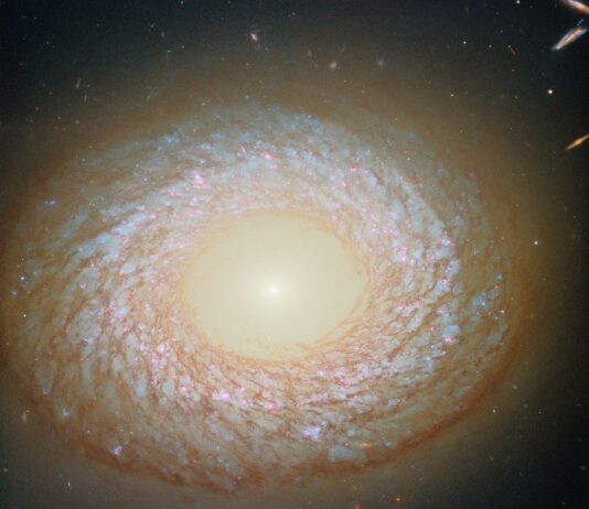 NASA’s Hubble Telescope Snaps Mysterious Galaxy Image A galaxy seen face-on, with a slightly elliptical disk that appears to have a hole in the center like a doughnut. In the hole, the core is a brightly glowing point that shines light out beyond the edge of the disk. Around the hole is an inner ring of dust, and at the galaxy’s edge is a thicker outer ring of dust, with a swirling web of dust strands in between. Blue stars and red nebulae are visible behind the dust.