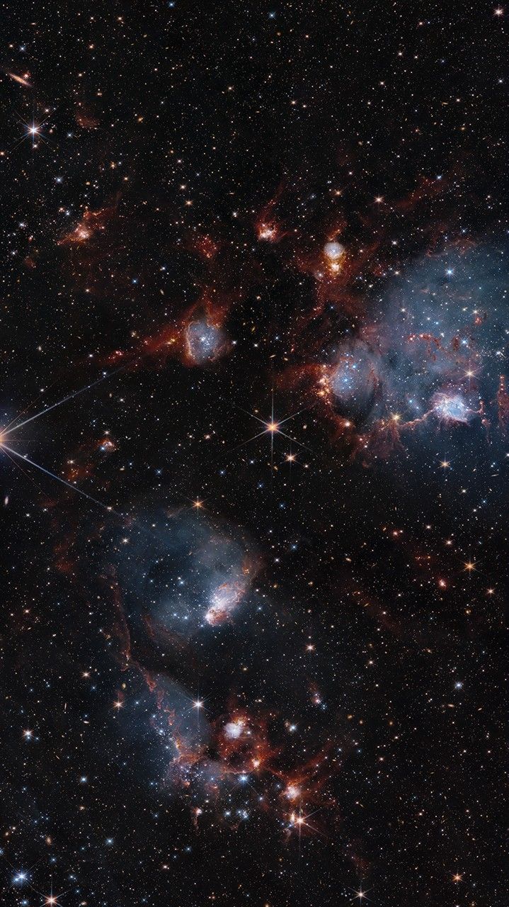 Bluish clouds of gas are wound through with red-glowing lanes and filaments of dust.. The clouds contain cavities filled with stars and many more stars are visible in the background and scattered throughout the clouds. The clouds are concentrated in the upper right and the lower middle of the image. Tiny background galaxies are also sprinkled throughout the image.