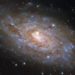 NASA’s Hubble Discovers Rare Cosmic Alignment A spiral galaxy, seen tilted at a slight angle, on a dark background of space. It glows softly from its center and out to its edge. The disk is a broad swirl of dark-reddish dust webs and sparkling blue patches where stars have formed. Atop the center of the galaxy is a foreground star that appears very large and bright with four diffraction spikes emanating from it. The star is relatively close to Earth compared to the galaxy.