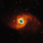 NASA’s Hubble Photographs a Star’s Final Performance The image holds a planetary nebula, a glowing shell of material thrown off by a dying star. A glowing, jagged ring encircles a small central region of greenish clouds, making it appear like a hole torn in fabric. This “hole” is itself encircled by a band of silvery-blue clouds and a larger, fainter, yellow ring of gas. Puffy, smoky clouds of orange and red gas billow out from there into a large oval nebula, fading into the dark background of space dotted by stars.