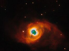 NASA’s Hubble Photographs a Star’s Final Performance The image holds a planetary nebula, a glowing shell of material thrown off by a dying star. A glowing, jagged ring encircles a small central region of greenish clouds, making it appear like a hole torn in fabric. This “hole” is itself encircled by a band of silvery-blue clouds and a larger, fainter, yellow ring of gas. Puffy, smoky clouds of orange and red gas billow out from there into a large oval nebula, fading into the dark background of space dotted by stars.
