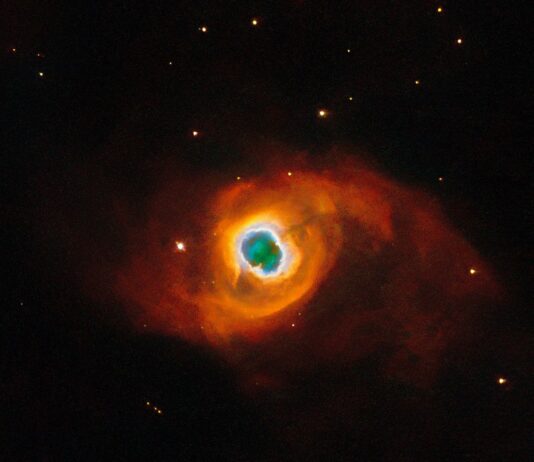 NASA’s Hubble Photographs a Star’s Final Performance The image holds a planetary nebula, a glowing shell of material thrown off by a dying star. A glowing, jagged ring encircles a small central region of greenish clouds, making it appear like a hole torn in fabric. This “hole” is itself encircled by a band of silvery-blue clouds and a larger, fainter, yellow ring of gas. Puffy, smoky clouds of orange and red gas billow out from there into a large oval nebula, fading into the dark background of space dotted by stars.