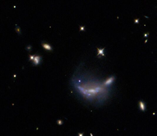 NASA’s Hubble Telescope Observes a Supernova Event In the exact center a supernova is visible as a small but bright blue dot. It lies atop the outer disk of a hazy-looking galaxy, which has a somewhat warped shape. Around this are a number of minor galaxies visible as glowing disks, and some points of light that are stars near to us, on a black background. A few bright stars hold X-shaped spikes that are optical artefacts from the telescope.