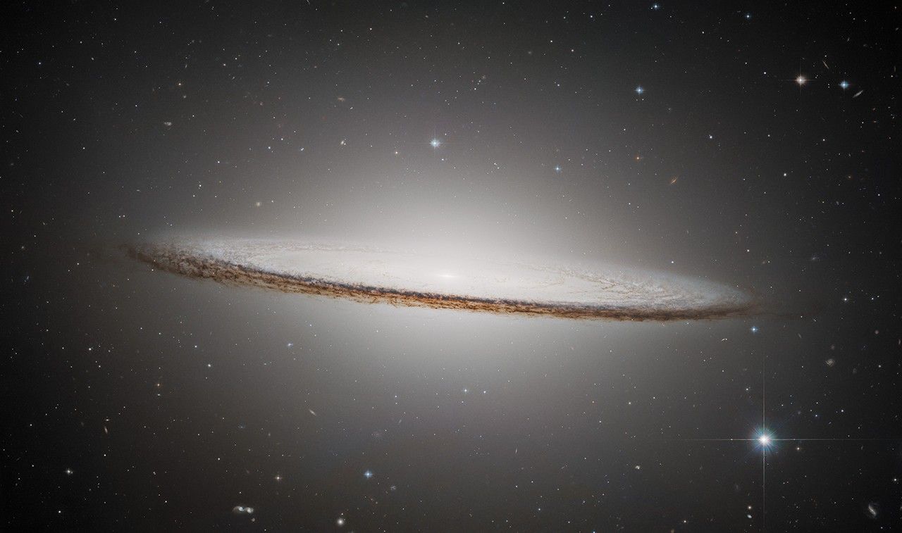 The Sombrero Galaxy is an oblong, pale white disk with a glowing core. It appears nearly edge-on but slants slightly in the front, presenting a slight top-down view of the inner region of the galaxy and its bright core. The outer disk is darker with shades of brown and black. Different colored distant galaxies and various stars are speckled among the black background of space surrounding the galaxy.