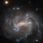 Hubble Detects Possible Imposter in Spiral Galaxy – NASA Science A spiral galaxy seen close to face-on. The center of its disk is a bright, pale yellowish, oval shape. Spiral arms extend from either side of the oval through the disk on irregular paths. The arms hold bright bluish-white patches of stars that mark their extent. Distant background galaxies appear as small orangish blobs around the spiral galaxy. In the top-left corner, a nearby star shines brightly, diffraction spikes are radiating from it.