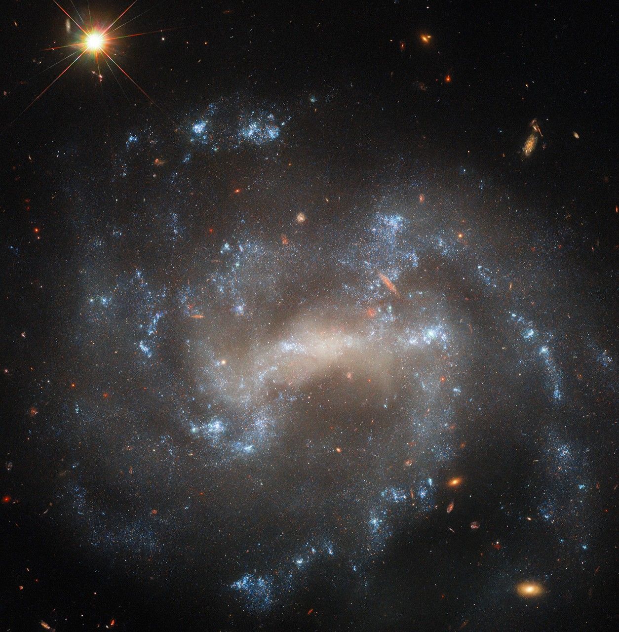 A spiral galaxy seen close to face-on. The center of its disk is a bright, pale yellowish, oval shape. Spiral arms extend from either side of the oval through the disk on irregular paths. The arms hold bright bluish-white patches of stars that mark their extent. Distant background galaxies appear as small orangish blobs around the spiral galaxy. In the top-left corner, a nearby star shines brightly, diffraction spikes are radiating from it.