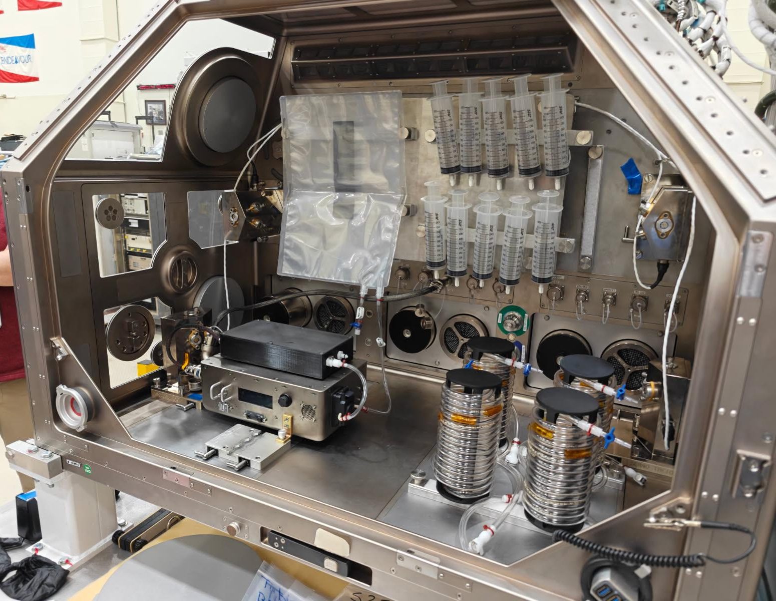 A large metal housing box containing the IVGEN Mini system is set up to create IV fluid. It includes the filtering system (two small boxes with cords attached), one plastic input bag, one plastic output bag, 10 syringes, additional cording, and four silver cylinder-shaped pieces of equipment.