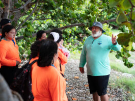 Interview with OCEANOS Instructor Samuel Suleiman – NASA A Puerto Rican man with a bushy white beard, green bucket hat, teal longsleeve shirt, and black shorts gestures to a group of high school students wearing neon orange shirts.