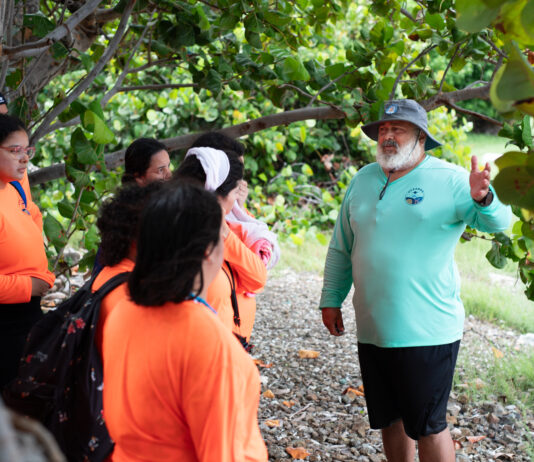 Chat with NASA’s OCEANOS Instructor Samuel Suleiman A Puerto Rican man with a bushy white beard, green bucket hat, teal longsleeve shirt, and black shorts gestures to a group of high school students wearing neon orange shirts.
