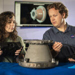 NASA’s Original Mission Revived Through Innovative Approaches Two NASA engineers examine a 3D-printed aluminum nozzle created under Marshall Space Flight Center’s Reactive Additive Manufacturing for the Fourth Industrial Revolution (RAMFIRE) project. One engineer holds a diagnostic device while the other inspects the nozzle’s surface. In the background, a computer screen displays a 3D model of the component, highlighting the project’s focus on innovation and advanced manufacturing techniques.