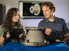 NASA’s Original Mission Revived Through Innovative Approaches Two NASA engineers examine a 3D-printed aluminum nozzle created under Marshall Space Flight Center’s Reactive Additive Manufacturing for the Fourth Industrial Revolution (RAMFIRE) project. One engineer holds a diagnostic device while the other inspects the nozzle’s surface. In the background, a computer screen displays a 3D model of the component, highlighting the project’s focus on innovation and advanced manufacturing techniques.