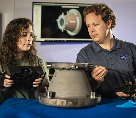 NASA’s Original Mission Revived Through Innovative Approaches Two NASA engineers examine a 3D-printed aluminum nozzle created under Marshall Space Flight Center’s Reactive Additive Manufacturing for the Fourth Industrial Revolution (RAMFIRE) project. One engineer holds a diagnostic device while the other inspects the nozzle’s surface. In the background, a computer screen displays a 3D model of the component, highlighting the project’s focus on innovation and advanced manufacturing techniques.