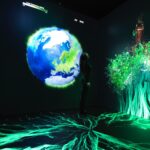 Media Preview of NASA’s New Earth Information Center A person stands in a dark room surrounded by images of green leaves and a large depiction of the Earth.