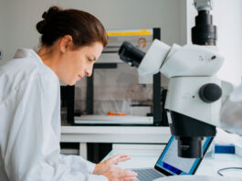 Institut Curie researchers gain time with new Copilot tool. A woman with her hair pulled back, wearing a white lab coat, works on a laptop in a laboratory, with a microscope in the foreground.