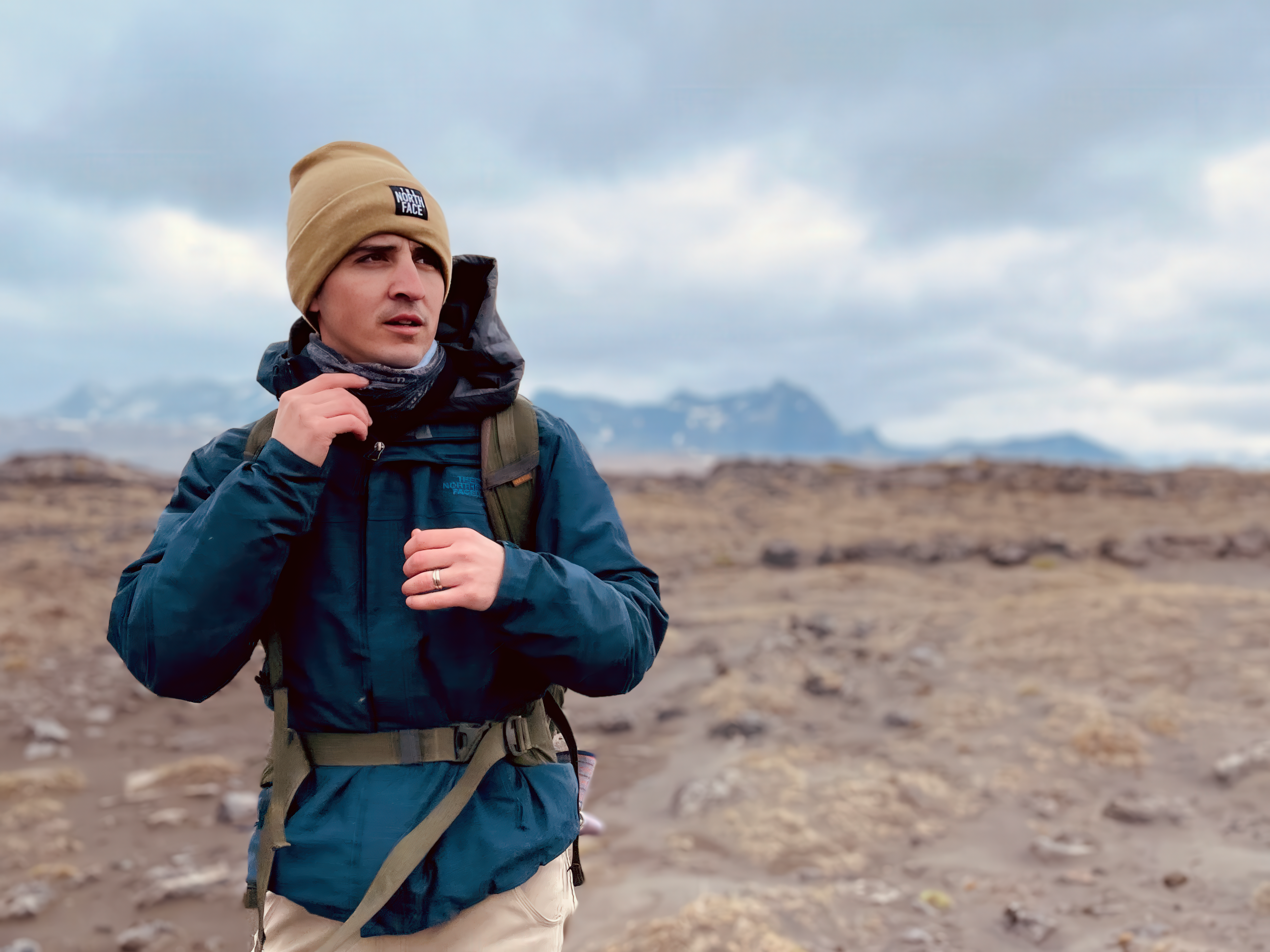 A man wearing a tan beanie and a blue winter jacket with a hiking backpack strapped around him. He