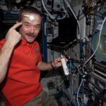 NASA Explores Space’s Impact on Mental Health and Well-Being Hadfield is wearing an orange polo shirt and khaki pants and holding a silver canister with a white label and blue tip in his left hand. With his right hand, he is pointing to a round yellow sensor taped to his forehead. There is a laptop over his left shoulder and multiple cords, wires, and switches on the wall in front of him.
