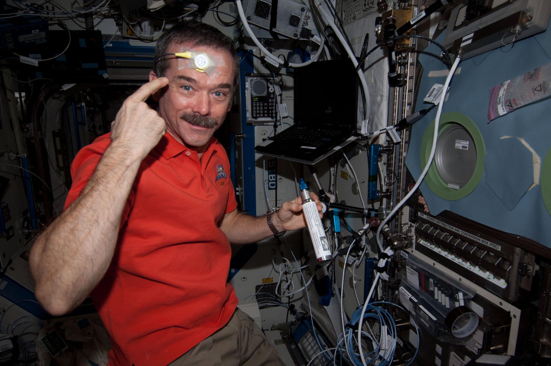 iss034e052585large.jpg Hadfield is wearing an orange polo shirt and khaki pants and holding a silver canister with a white label and blue tip in his left hand. With his right hand, he is pointing to a round yellow sensor taped to his forehead. There is a laptop over his left shoulder and multiple cords, wires, and switches on the wall in front of him.