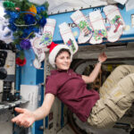NASA: Festive Season Celebrations in Space A woman with short, dark hair and wearing a Santa hat, maroon shirt, and khaki pants smiles with arms outspread while floating in microgravity in front of Christmas stockings.