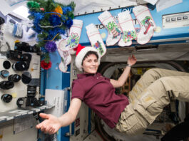 NASA: Festive Season Celebrations in Space A woman with short, dark hair and wearing a Santa hat, maroon shirt, and khaki pants smiles with arms outspread while floating in microgravity in front of Christmas stockings.