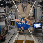 NASA Achieves Optimal Performance in Zero Gravity Saint-Jacques is wearing a blue polo shirt with a CSA logo, khaki pants, and black socks. He is suspended upside down by four white straps connected to a harness around his middle and has a virtual reality headset on his head and a small black controller in his right hand. An open laptop floats next to him.