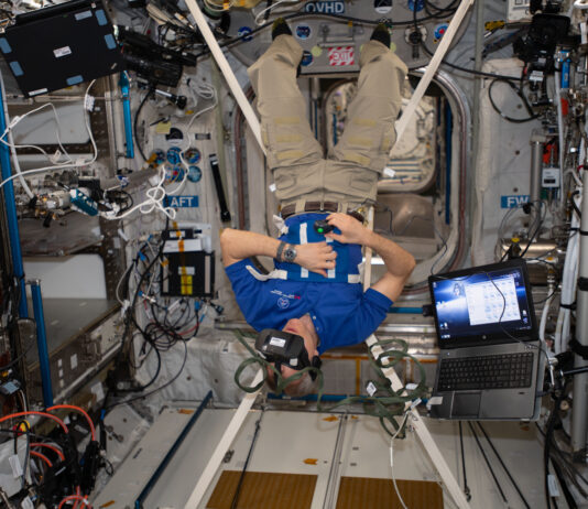 NASA Achieves Optimal Performance in Zero Gravity Saint-Jacques is wearing a blue polo shirt with a CSA logo, khaki pants, and black socks. He is suspended upside down by four white straps connected to a harness around his middle and has a virtual reality headset on his head and a small black controller in his right hand. An open laptop floats next to him.