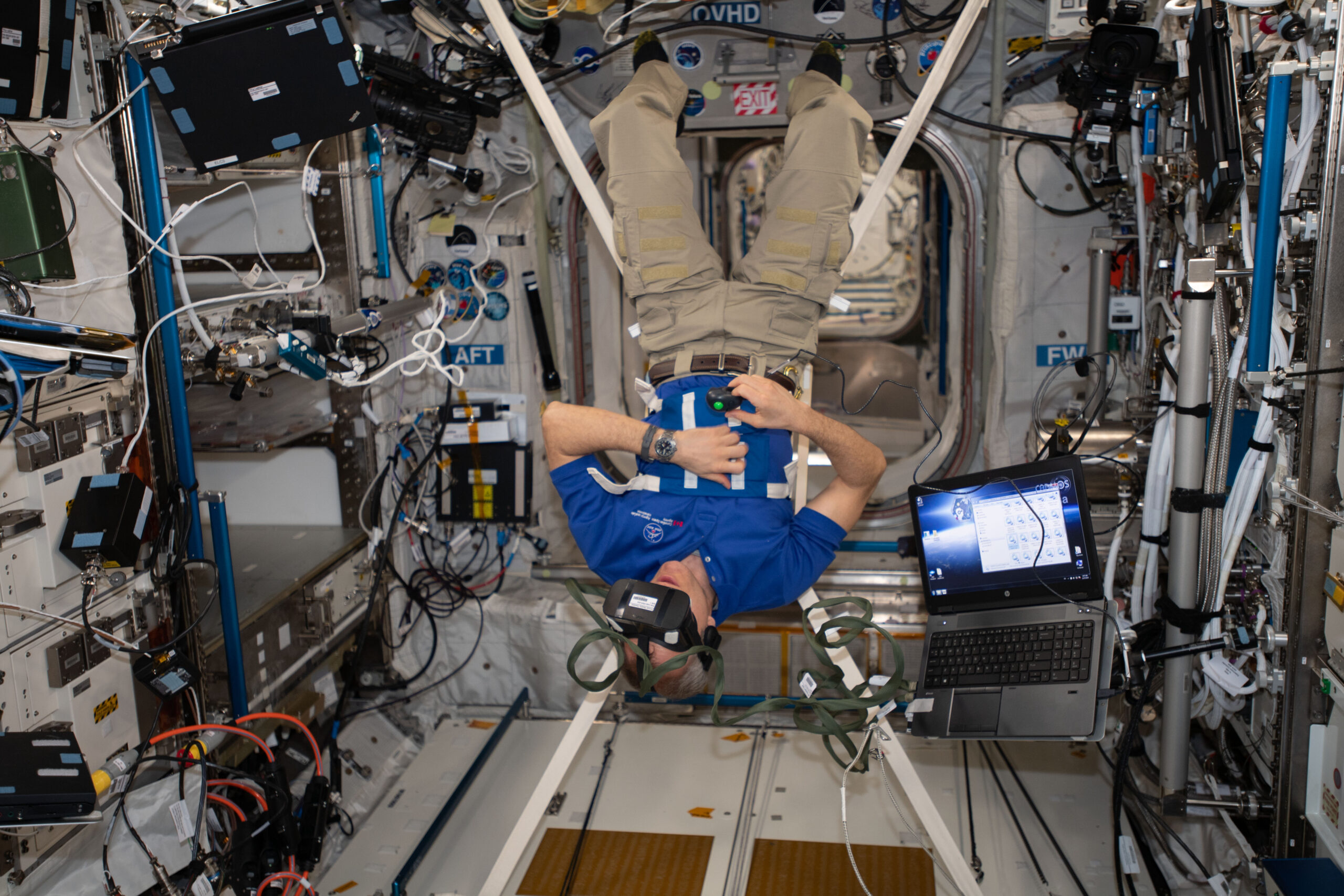 Saint-Jacques is wearing a blue polo shirt with a CSA logo, khaki pants, and black socks. He is suspended upside down by four white straps connected to a harness around his middle and has a virtual reality headset on his head and a small black controller in his right hand. An open laptop floats next to him.