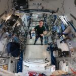 NASA’s Advances in Navigation Technology Maurer stands in the middle of this circular fisheye image, facing the camera, wearing a green t-shirt and black pants. In his right hand, he holds a cube-shaped Astrobee, which has black sides and a white panel facing the camera with two bright blue lights that look sort of like eyes. Maurer is pointing at the Astrobee with his left hand.