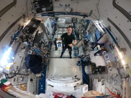NASA’s Advances in Navigation Technology Maurer stands in the middle of this circular fisheye image, facing the camera, wearing a green t-shirt and black pants. In his right hand, he holds a cube-shaped Astrobee, which has black sides and a white panel facing the camera with two bright blue lights that look sort of like eyes. Maurer is pointing at the Astrobee with his left hand.