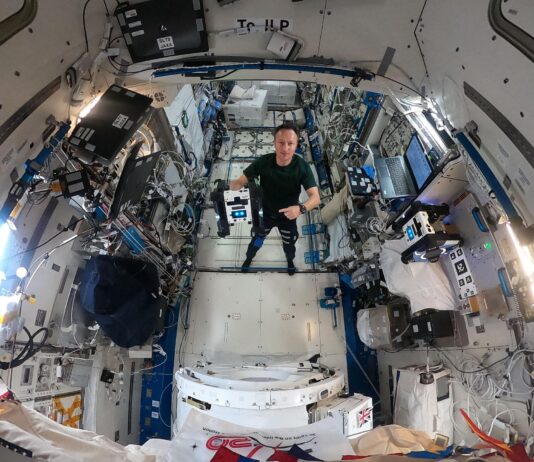 NASA’s Advances in Navigation Technology Maurer stands in the middle of this circular fisheye image, facing the camera, wearing a green t-shirt and black pants. In his right hand, he holds a cube-shaped Astrobee, which has black sides and a white panel facing the camera with two bright blue lights that look sort of like eyes. Maurer is pointing at the Astrobee with his left hand.