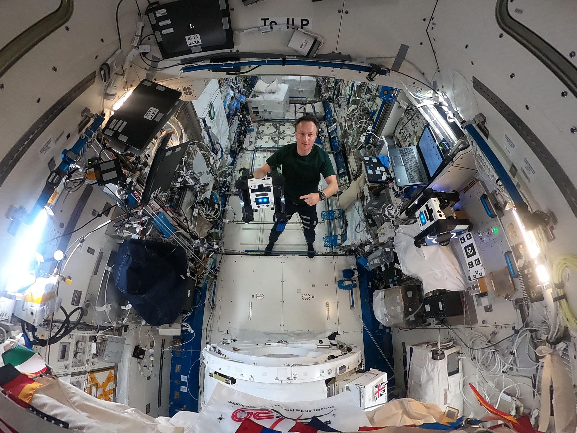 iss066e087003large.jpg Maurer stands in the middle of this circular fisheye image, facing the camera, wearing a green t-shirt and black pants. In his right hand, he holds a cube-shaped Astrobee, which has black sides and a white panel facing the camera with two bright blue lights that look sort of like eyes. Maurer is pointing at the Astrobee with his left hand.