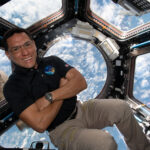 Astronaut Frank Rubio Observes Earth from Space Station Cupola A man in a dark polo shirt smiles at the camera with his arms crossed. He is in the cupola of the International Space Station, an area with multiple windows through which Earth and space can be seen. Earth