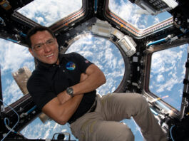 Astronaut Frank Rubio Observes Earth from Space Station Cupola A man in a dark polo shirt smiles at the camera with his arms crossed. He is in the cupola of the International Space Station, an area with multiple windows through which Earth and space can be seen. Earth