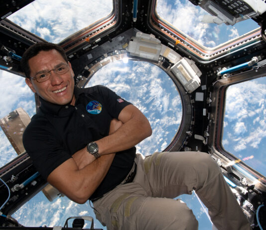 Astronaut Frank Rubio Observes Earth from Space Station Cupola A man in a dark polo shirt smiles at the camera with his arms crossed. He is in the cupola of the International Space Station, an area with multiple windows through which Earth and space can be seen. Earth
