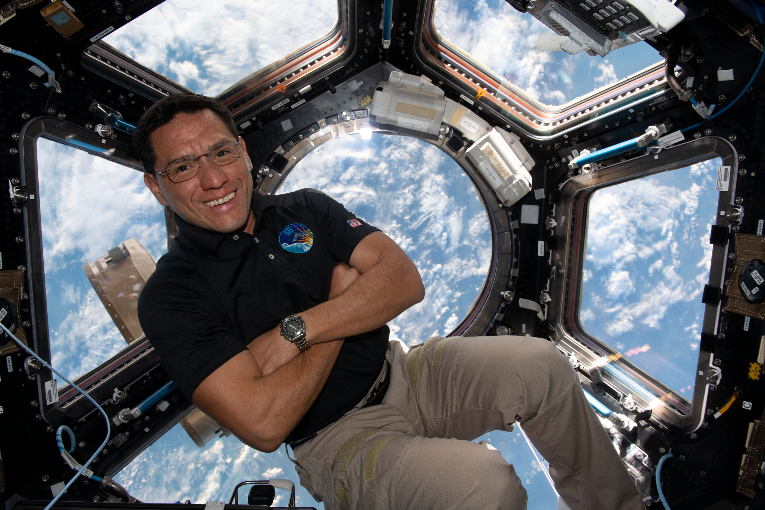 A man in a dark polo shirt smiles at the camera with his arms crossed. He is in the cupola of the International Space Station, an area with multiple windows through which Earth and space can be seen. Earth