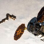 Northrop Grumman, SpaceX Falcon 9 Launch Coverage Announced by NASA A cylinder-shaped spacecraft with prominent cymbal-shaped solar arrays on either side of it nears a long robotic arm, with a cloudy Earth below.