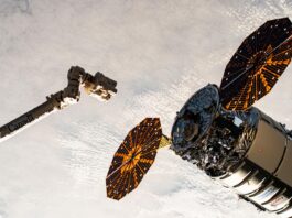 Northrop Grumman, SpaceX Falcon 9 Launch Coverage Announced by NASA A cylinder-shaped spacecraft with prominent cymbal-shaped solar arrays on either side of it nears a long robotic arm, with a cloudy Earth below.