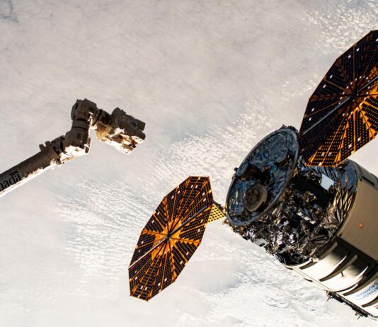 Northrop Grumman, SpaceX Falcon 9 Launch Coverage Announced by NASA A cylinder-shaped spacecraft with prominent cymbal-shaped solar arrays on either side of it nears a long robotic arm, with a cloudy Earth below.
