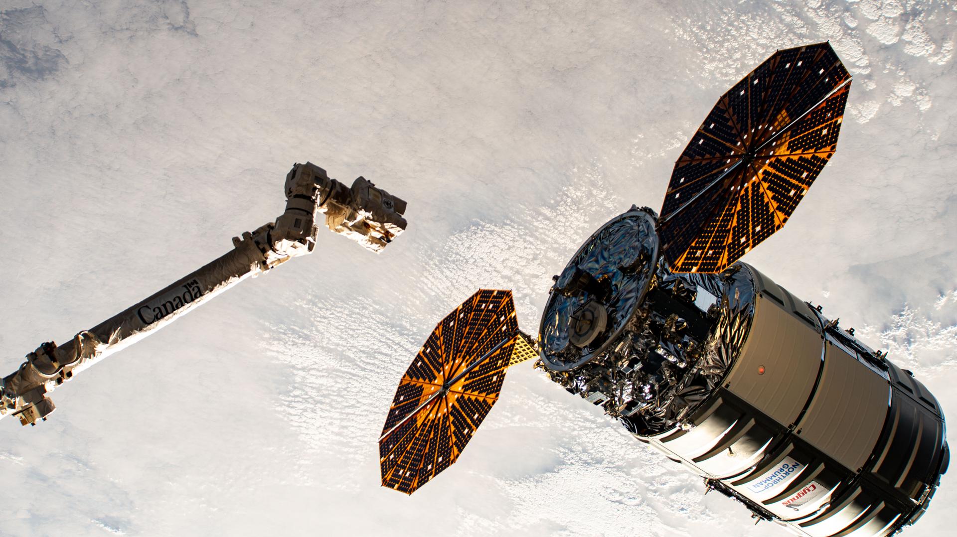 A cylinder-shaped spacecraft with prominent cymbal-shaped solar arrays on either side of it nears a long robotic arm, with a cloudy Earth below.