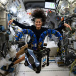 Suni Williams Collaborates with NASA’s Astrobee in Space Mission Astrobee, a robot that looks like a small black cube with two blue tentacle-like arms on top. Behind it, astronaut Suni Williams stretches her arms out to match Astrobee