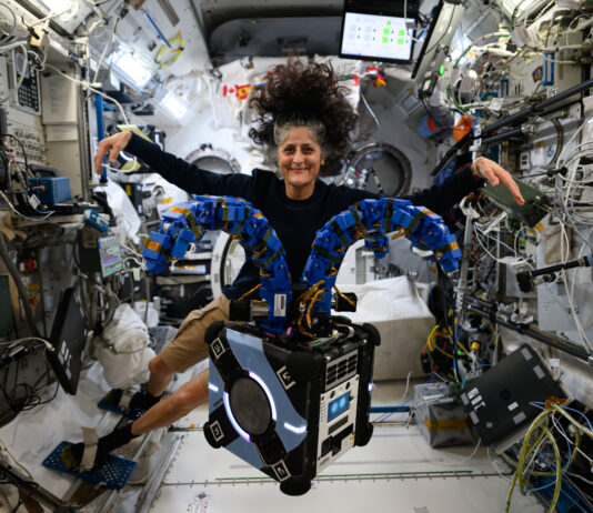 Suni Williams Collaborates with NASA’s Astrobee in Space Mission Astrobee, a robot that looks like a small black cube with two blue tentacle-like arms on top. Behind it, astronaut Suni Williams stretches her arms out to match Astrobee