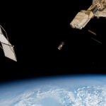 JAXA Launches Wooden Satellite from Space Station with NASA An outside view of the space station with three square boxes in the center of the image. To the left is a solar panel. To the right is part of the space station. At the bottom is a view of the Earth.