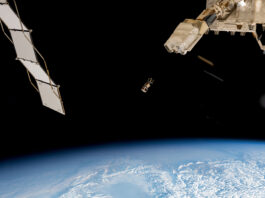 JAXA Launches Wooden Satellite from Space Station with NASA An outside view of the space station with three square boxes in the center of the image. To the left is a solar panel. To the right is part of the space station. At the bottom is a view of the Earth.