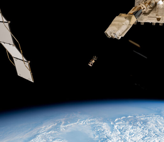 JAXA Launches Wooden Satellite from Space Station with NASA An outside view of the space station with three square boxes in the center of the image. To the left is a solar panel. To the right is part of the space station. At the bottom is a view of the Earth.