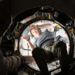 NASA’s Unique Approach to Space Operations We look upward at an Asian man holding a silver tablet. He is wearing a gray t-shirt and green pants with a belt. He is halfway through a round opening. The inside of the craft he