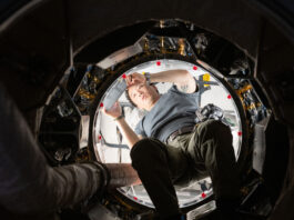 NASA’s Unique Approach to Space Operations We look upward at an Asian man holding a silver tablet. He is wearing a gray t-shirt and green pants with a belt. He is halfway through a round opening. The inside of the craft he
